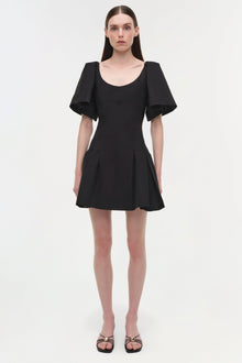 Black mini dress with puffed short sleeves, scoop neckline, and pleated skirt, worn with black strappy sandals.