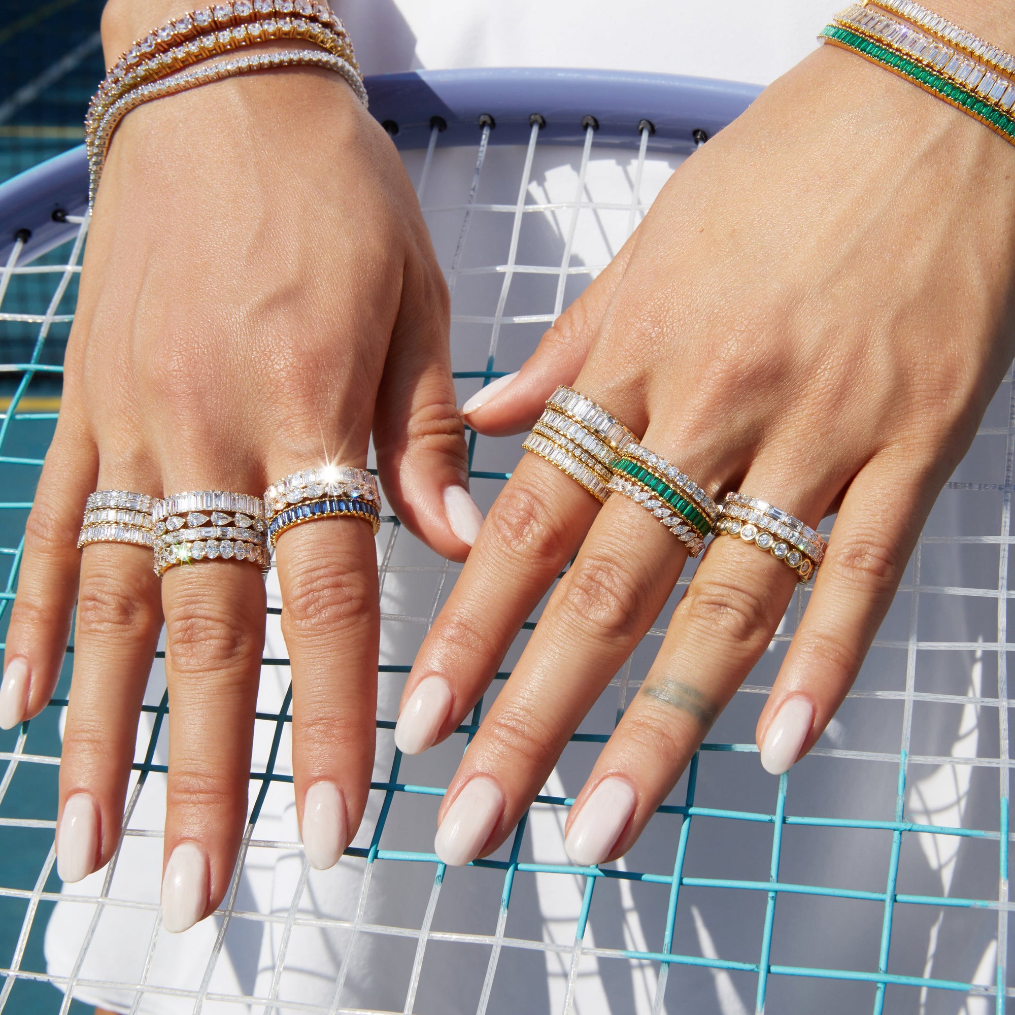 Lifestyle Model Close-up of a woman's hands resting on a tennis racket, wearing multiple stacked gold rings and bracelets adorned with diamonds, green stones, and blue stones