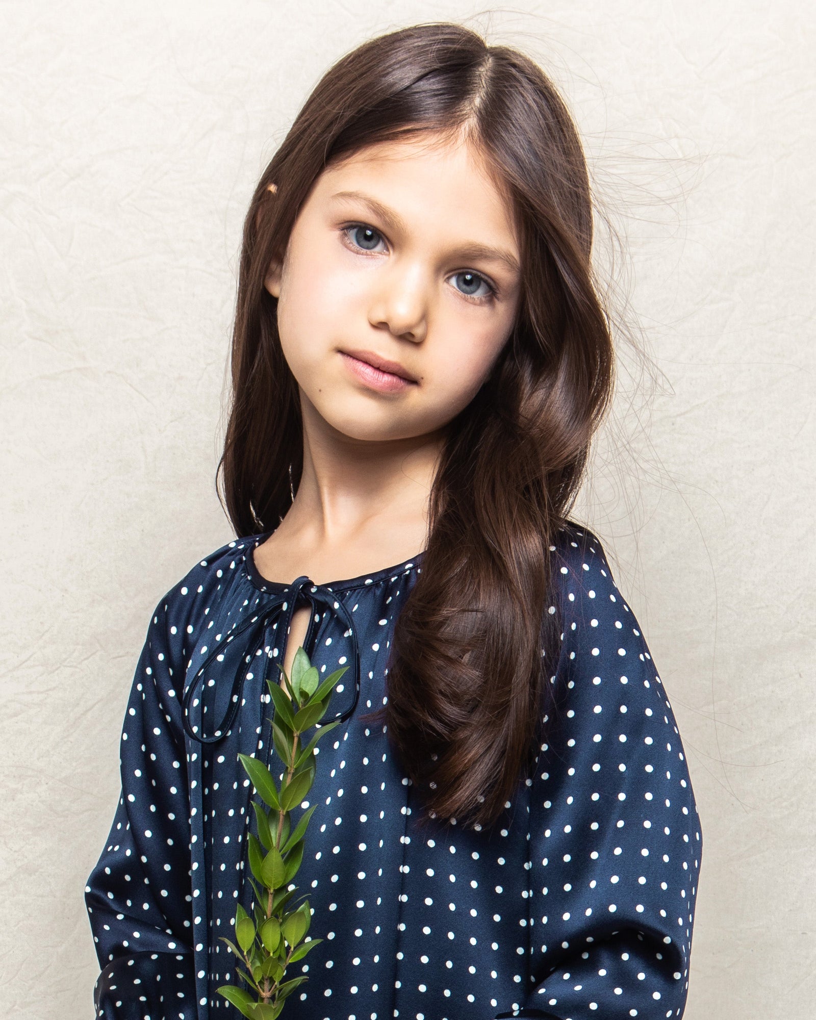 A girl with long brown hair and blue eyes in a Petite Plume Girls Silk Delphine Nightgown in Navy Polka Dot holds a green leafy stem against a light gray background, gazing softly at the camera, her serene presence evoking the elegance of luxurious sleepwear.