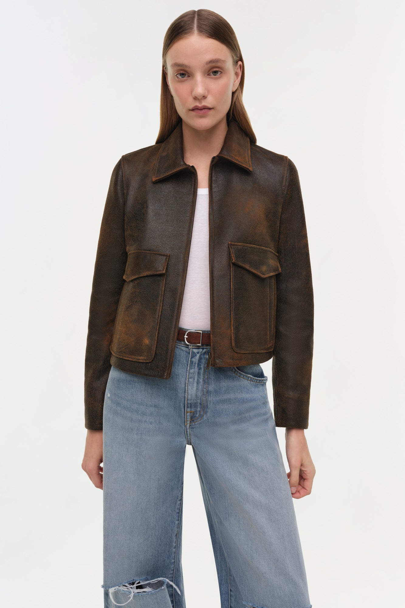 Brown leather jacket with spread collar, front zip closure, and two flap chest pockets worn over white tee and light blue jea