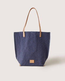 The graf lantz Hana Canvas Tote is a stylish blue denim bag made from durable cotton canvas, featuring tan leather handles and a small tan leather patch on the front, set against a plain light background.