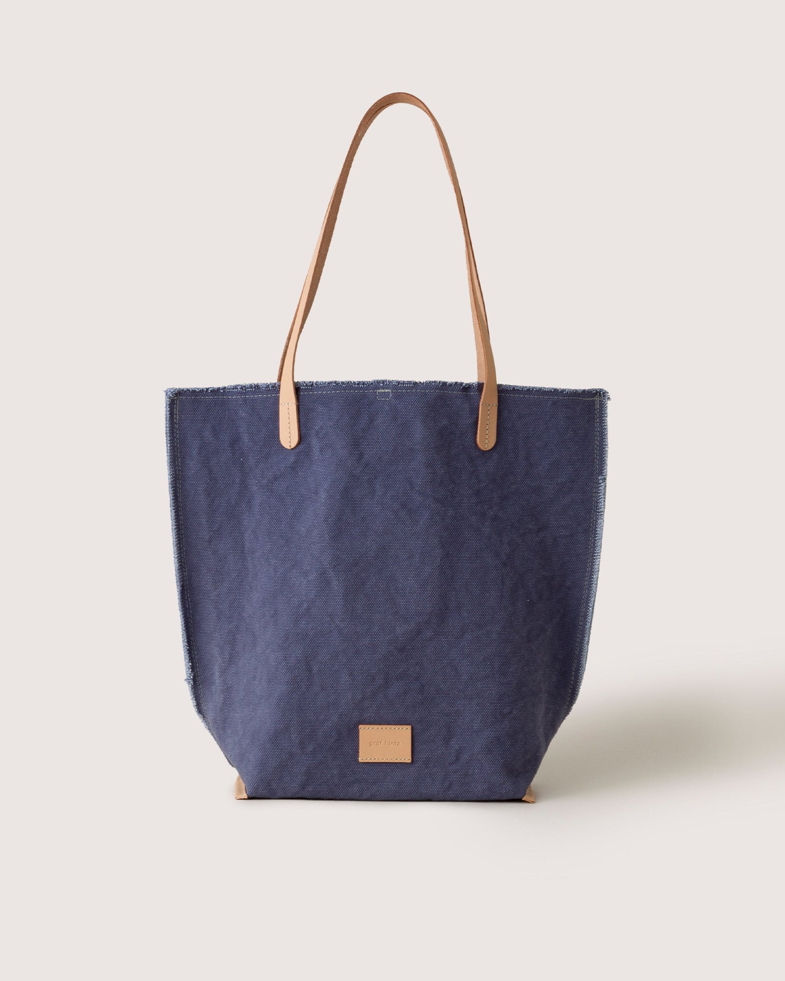 The graf lantz Hana Canvas Tote is a stylish blue denim bag made from durable cotton canvas, featuring tan leather handles and a small tan leather patch on the front, set against a plain light background.