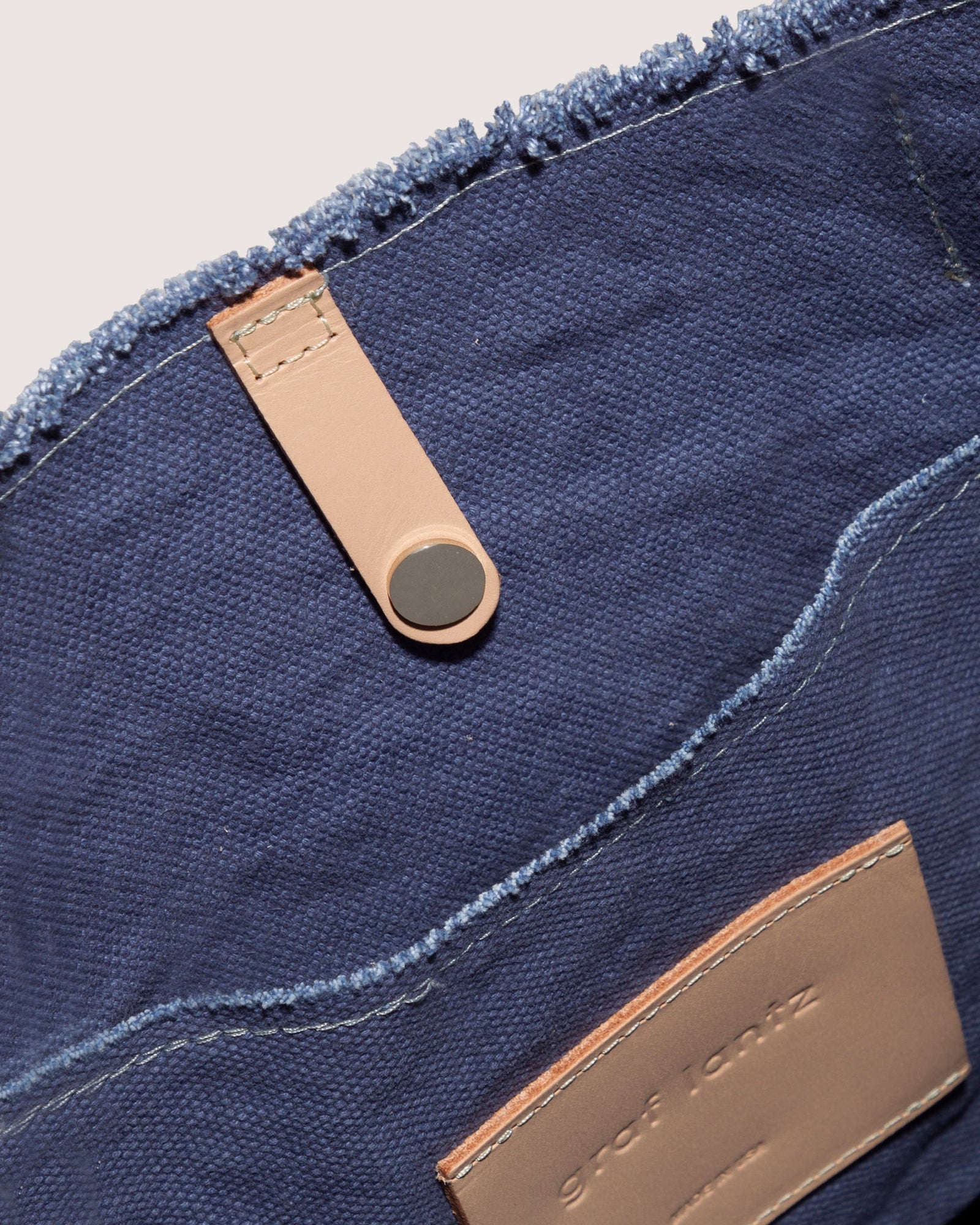 Close-up of a blue textile bag by graf lantz, made from durable cotton canvas with frayed edges. Inside, the Hana Canvas Tote features a tan leather strap with black snap button and textured tan patch with visible stitches. The fabric looks soft and sturdy.