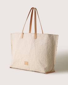 A large beige canvas tote bag by graf lantz, featuring light brown vegetable-tanned leather handles and a small rectangular patch on the front, is set against a plain off-white background.