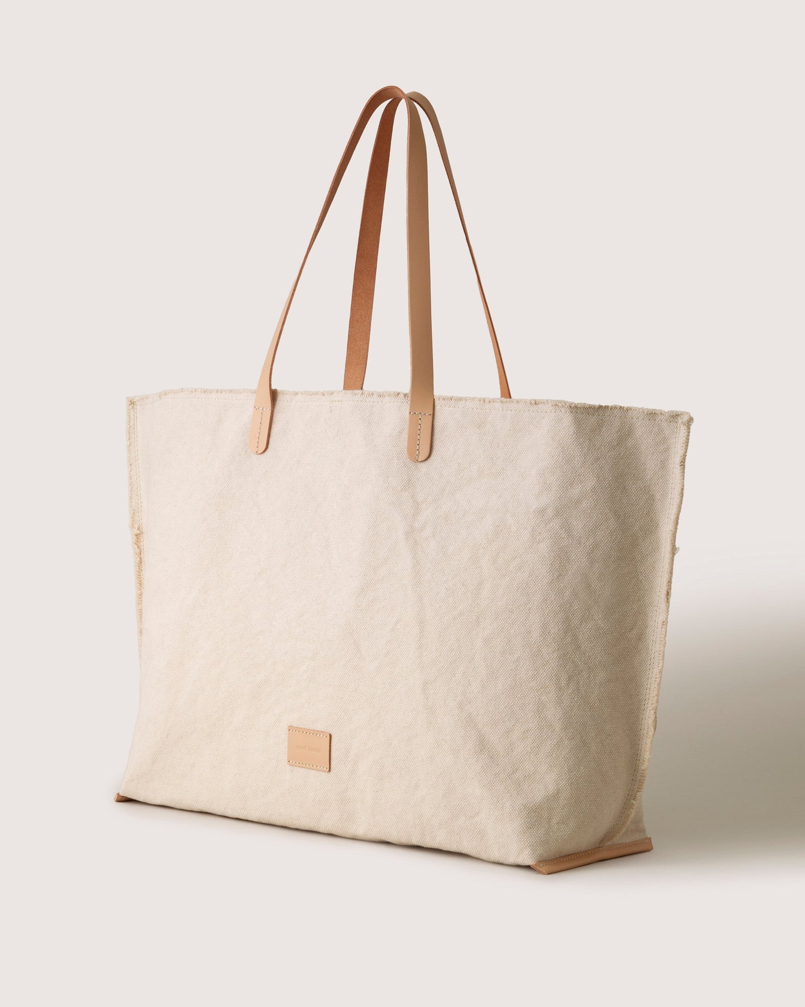 A large beige canvas tote bag by graf lantz, featuring light brown vegetable-tanned leather handles and a small rectangular patch on the front, is set against a plain off-white background.