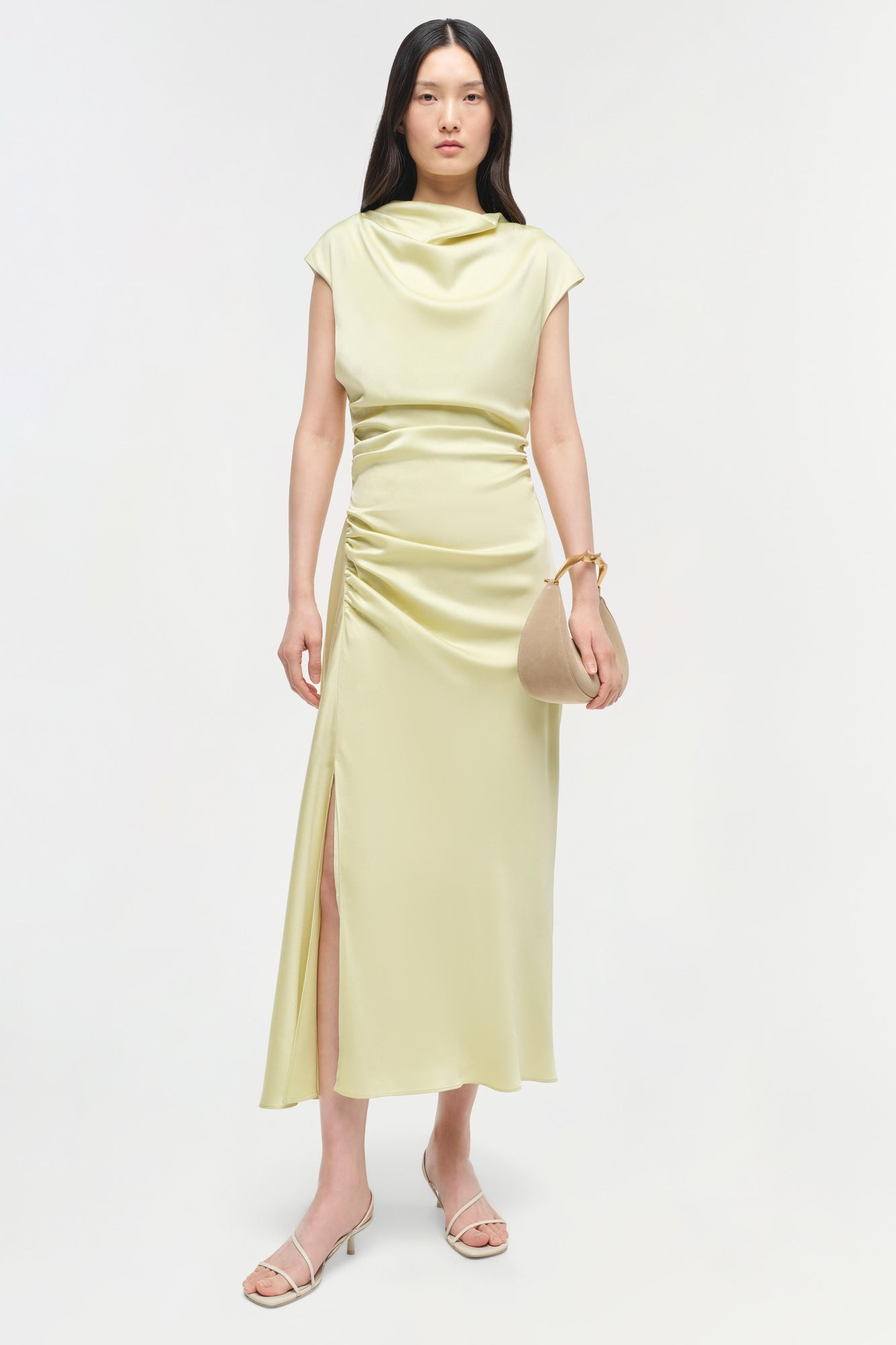 Pale yellow satin midi dress with cowl neckline, ruched waist, and side slit, worn with white strappy sandals.