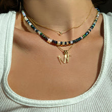 Shimmering beaded necklace made of faceted opals in shades of fiery pastels, grey, teal, and black on a gold linking ovals clasp. Styled on a neck layered with a stitch necklace, bead chain necklace, M and T letter charms.