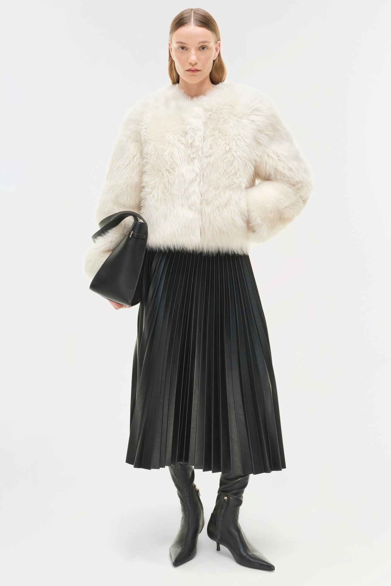 Woman wearing cream-colored shearling jacket with black pleated skirt and black boots, holding a black handbag.