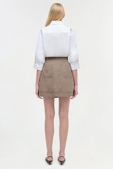 Model wearing white collared shirt with three-quarter sleeves over beige mini skirt with patch pockets, viewed from back.