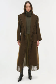 Olive wool coat with fringe trim along the front and hem, worn over a turtleneck sweater.