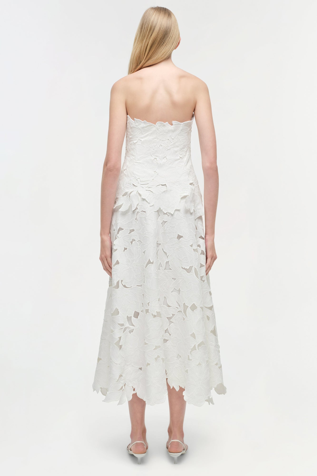 White strapless midi dress with leaf-shaped embroidered cutout details throughout, scalloped hemline, worn with clear heeled 