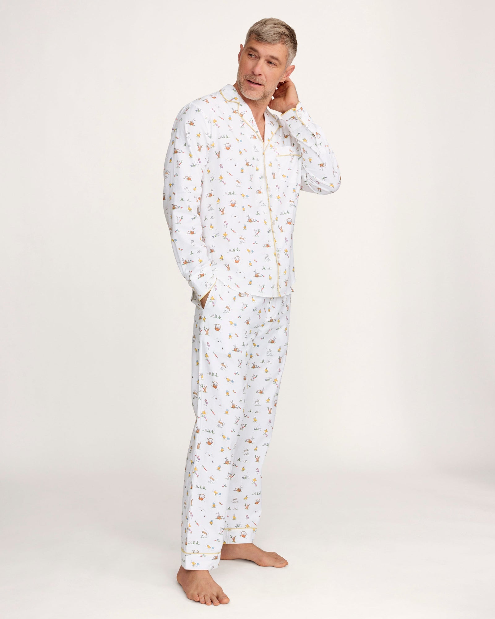 A man with short gray hair wears the Men's Twill Pajama Set in Easter Frolic—white pants and a long-sleeve top with a small, colorful print—standing barefoot against a plain light background.