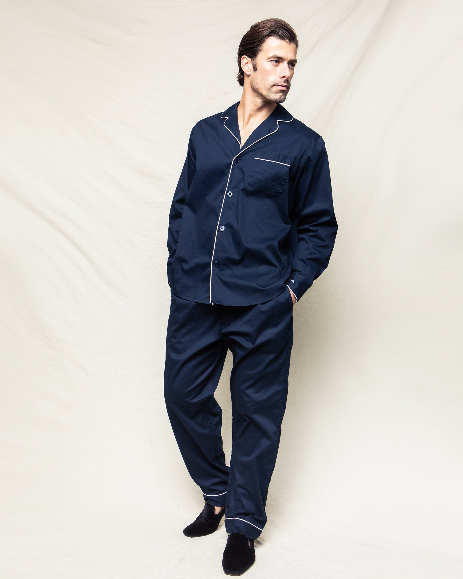 A man wearing Petite Plumes Mens Twill Pajamas in Navy with White Piping stands against a light background, hands in pockets and legs slightly crossed. He looks left while wearing black slippers.