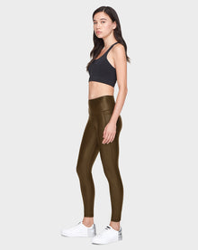 A woman in profile poses against a plain backdrop, wearing Rudsaks SHIRIN L - 8521034 Army shiny olive green vegan leather leggings with a black sports bra and white sneakers. She has long dark hair and a neutral expression.