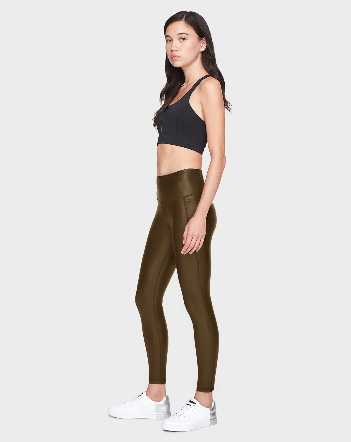 A woman in profile poses against a plain backdrop, wearing Rudsaks SHIRIN L - 8521034 Army shiny olive green vegan leather leggings with a black sports bra and white sneakers. She has long dark hair and a neutral expression.