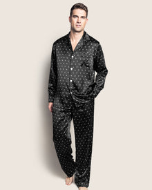 A man barefoot against a light background wears Petite Plumes Mens Silk Pajama Set in Black Art Nouveau. The elegant set, with a button-up shirt and matching pants, mirrors the luxury of mulberry silk. One hand in his pocket, he gazes slightly upward.