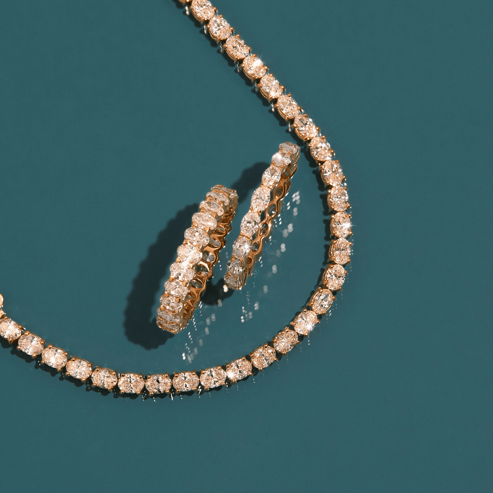 Flatlay Model A gold diamond tennis necklace and two gold diamond hoop earrings resting on a dark teal background.
