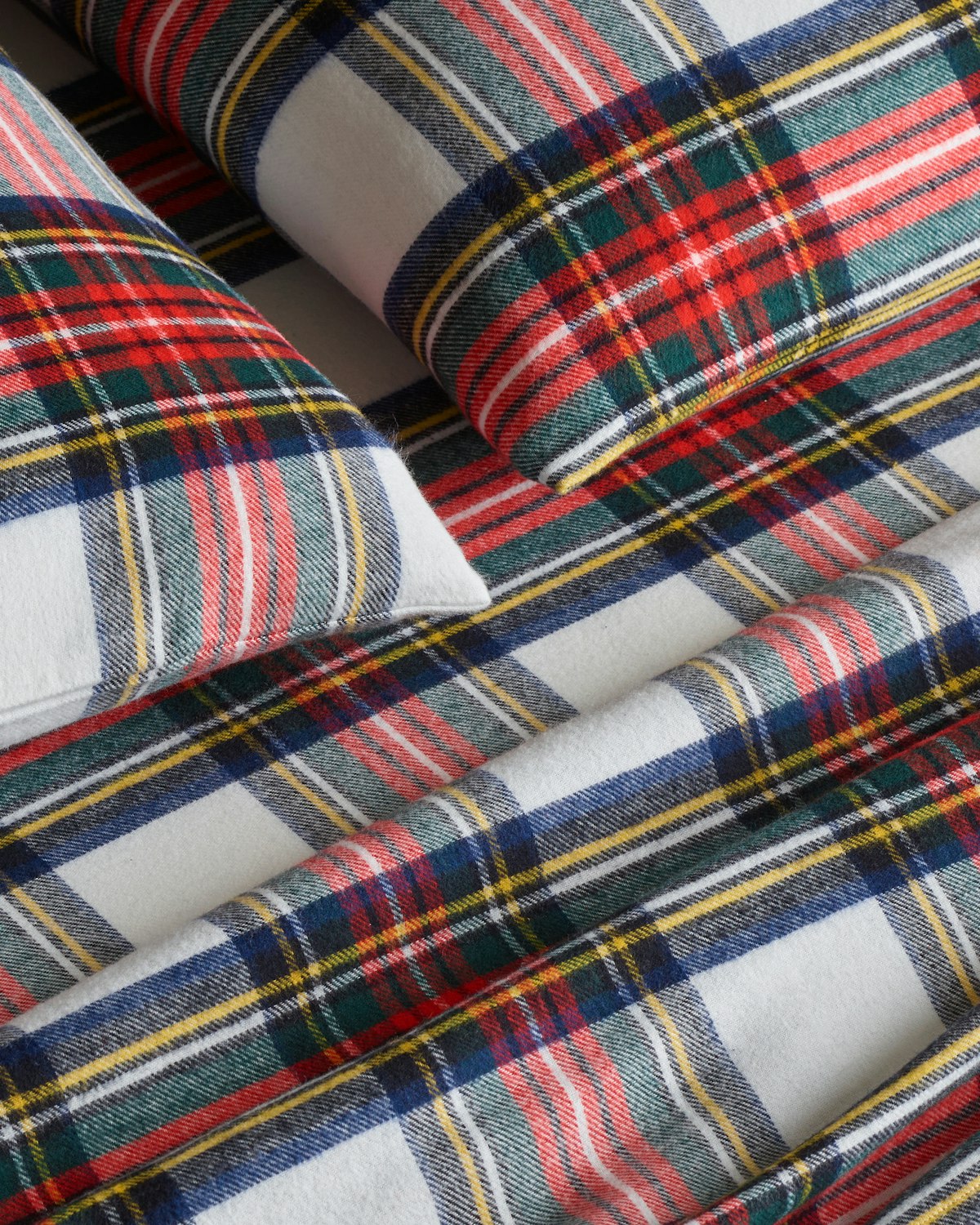 Close-up of a bed adorned with Petite Plumes Flannel Sheets in Balmoral Tartan, showcasing plaid sheets and pillowcases accented with red, green, blue, and yellow stripes on white. The cozy cotton brushed flannel creates a warm and inviting atmosphere.