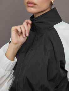 Model wears a cropped windbreaker with a popped collar, black bodice and white long sleeves with a cinched wristline.