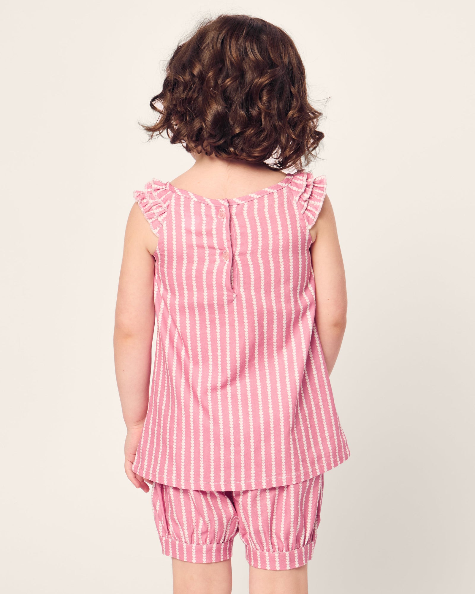 A young child with curly hair faces away, wearing Petite Plumes Girls Twill Amelie Short Set in Love Lines. The pink and white striped sleeveless top features ruffled shoulders and matching shorts, crafted from soft cotton sleepwear material, set against an off-white background.