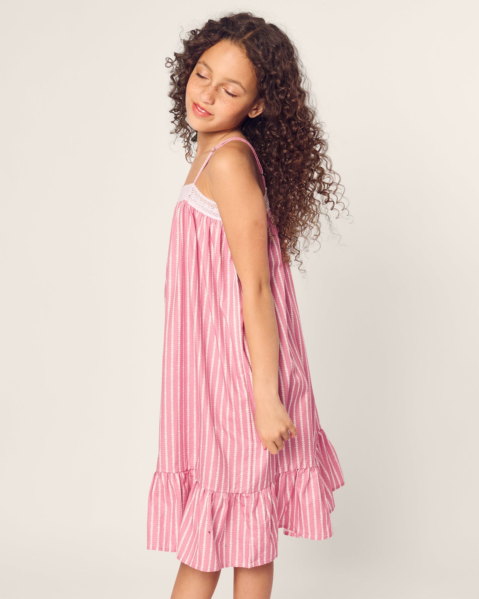 The girl with curly hair, wearing a Girls Twill Lilly Nightgown in Love Lines by Petite Plume, stands serenely with eyes closed against a light background.