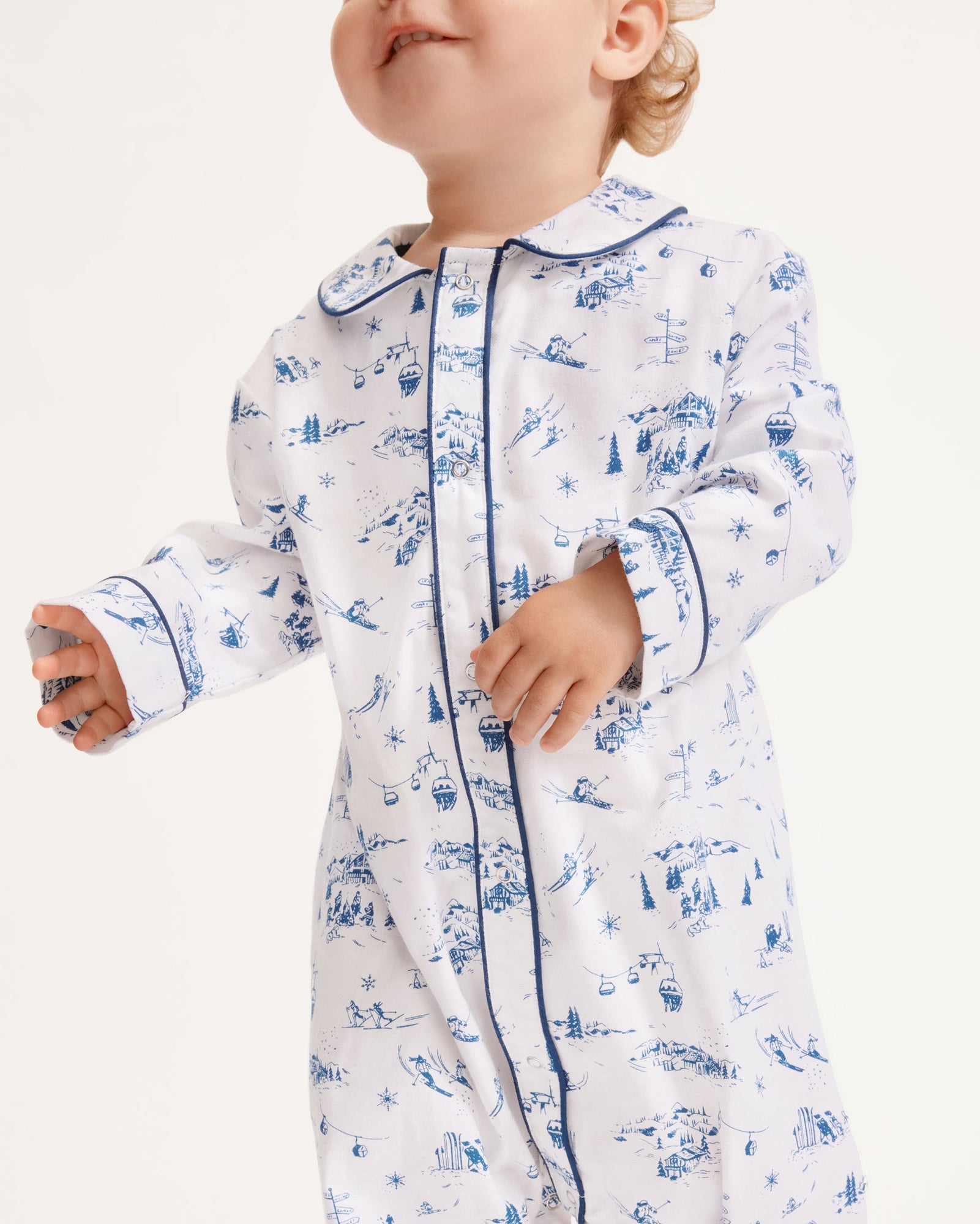 The Baby's Twill Cambridge Romper in Chalet Toile is a white long-sleeve, full-length onesie with blue alpine scene prints and blue piping on the collar, cuffs, and front placket.