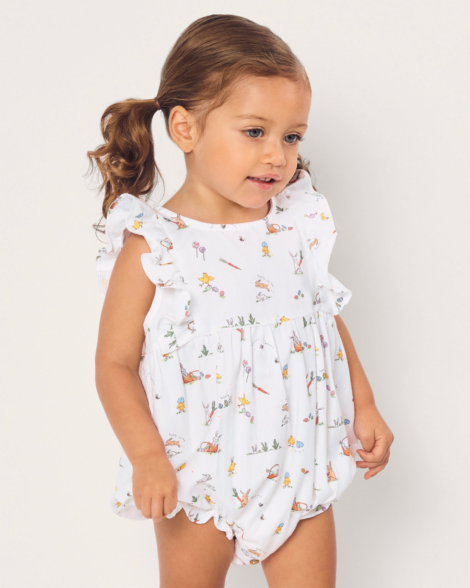 A young child with brown hair in pigtails wears the Baby's Twill Ruffled Romper in Easter Frolic, featuring colorful animal and floral prints, standing against a plain light background.