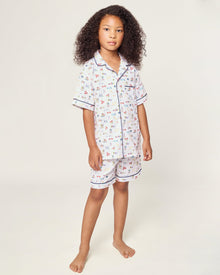 The Kid's Twill Short Set in Literary Lessons features a white base with colorful school-themed prints—pencils, books, and erasers—plus navy piping on the collar, pocket, sleeves, and shorts.
