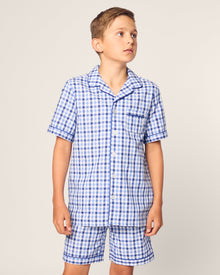 A young boy models the Kids Twill Pajama Short Set in Royal Blue Gingham by Petite Plume. The set features a yarn-dyed cotton short-sleeved button-up shirt with a collar and chest pocket, paired with shorts, as he gazes to the side against a plain backdrop.