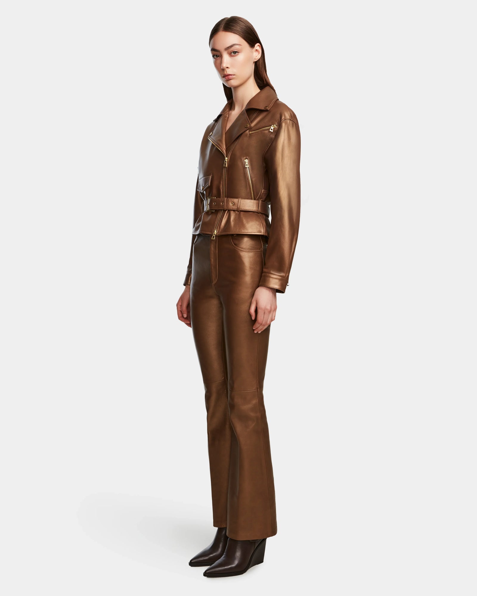 A woman stands against a plain background wearing the Rudsak ARIELLE - 8124052 Antique Bronze lamb leather biker jacket with matching belt and brown leather pants, paired with dark ankle boots.