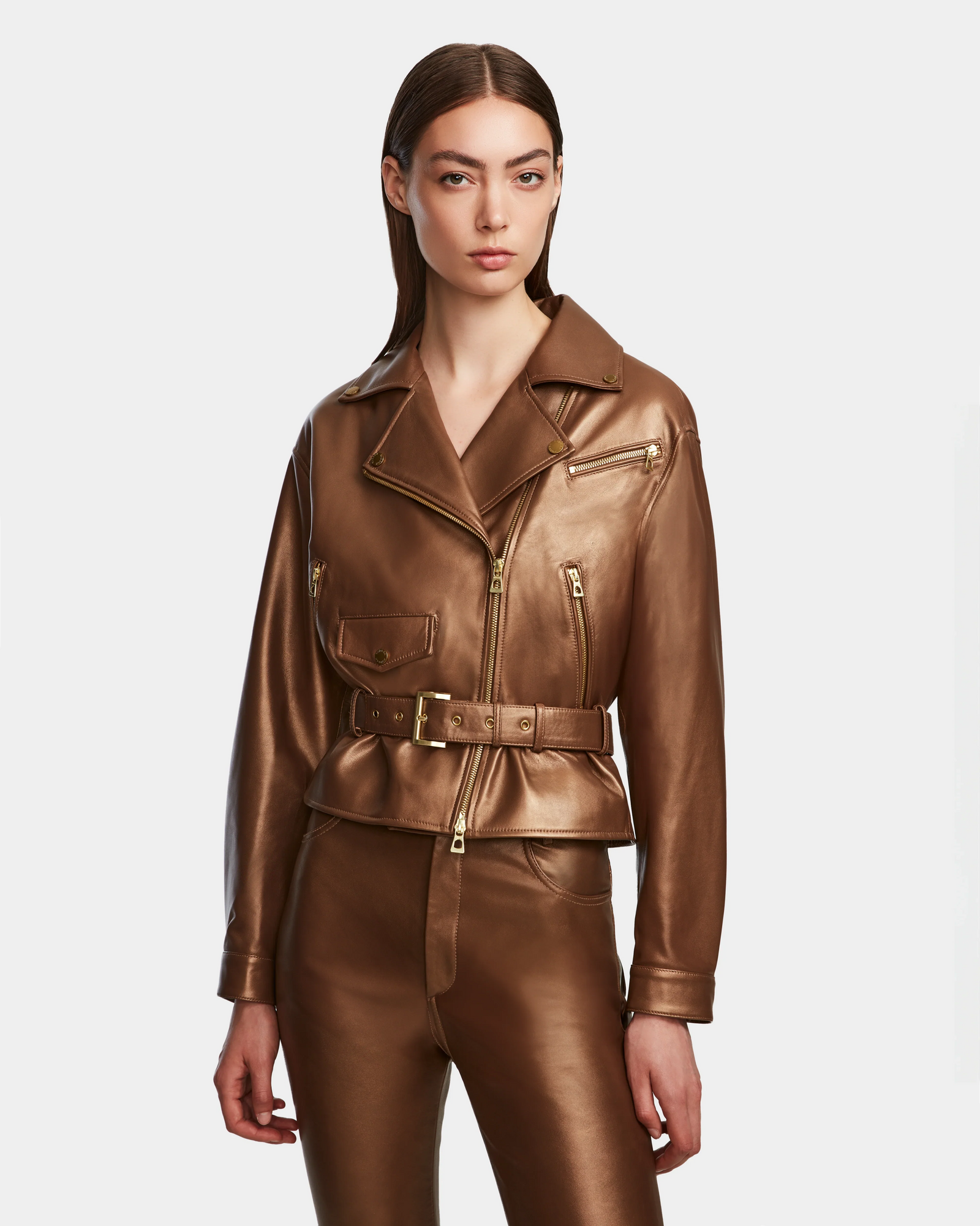 A woman wearing the Rudsak ARIELLE - 8124052 Antique Bronze shiny brown lamb leather jacket with gold zippers and matching brown leather pants, standing against a plain white background.