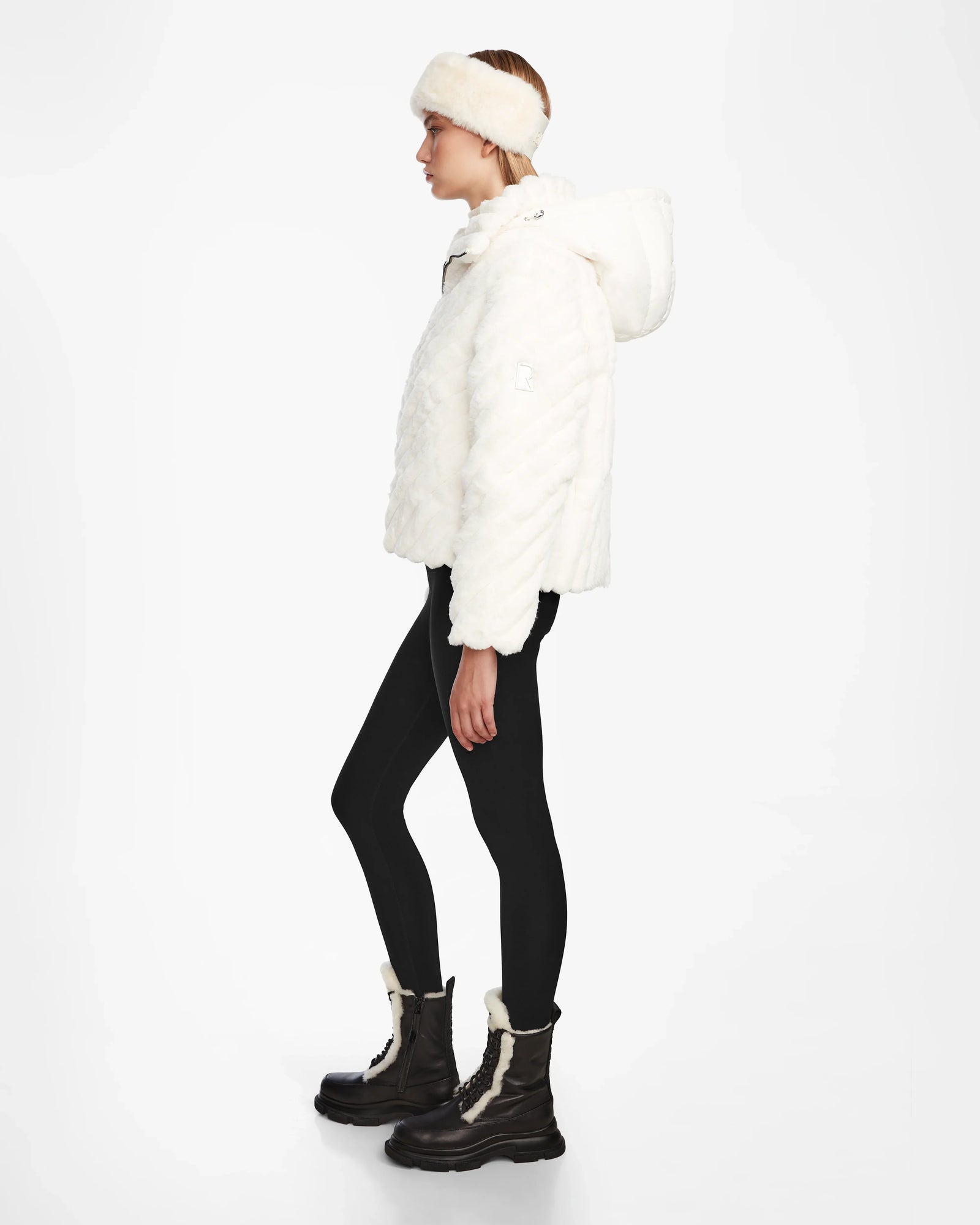 A person in profile wears the Rudsak ANASTASIA - 8124804 Iceland white velvet puffer jacket with a matching faux fur headband, black leggings, and tall dark lace-up boots with cream accents, set against a plain white background.