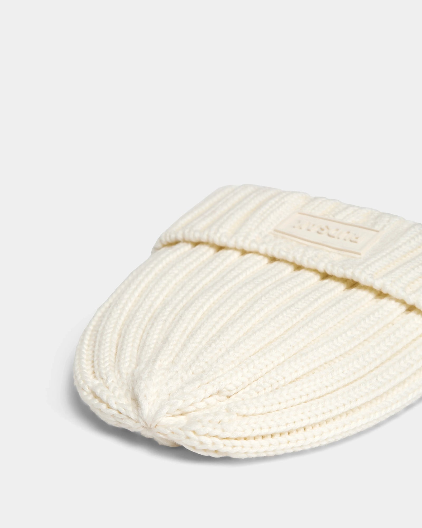 A close-up of the Rudsak ANISA - L423500 Optic White ribbed knit beanie with a rectangular patch on the turned-up brim, perfect for elevating your winter wardrobe. Shown folded on a plain light background.
