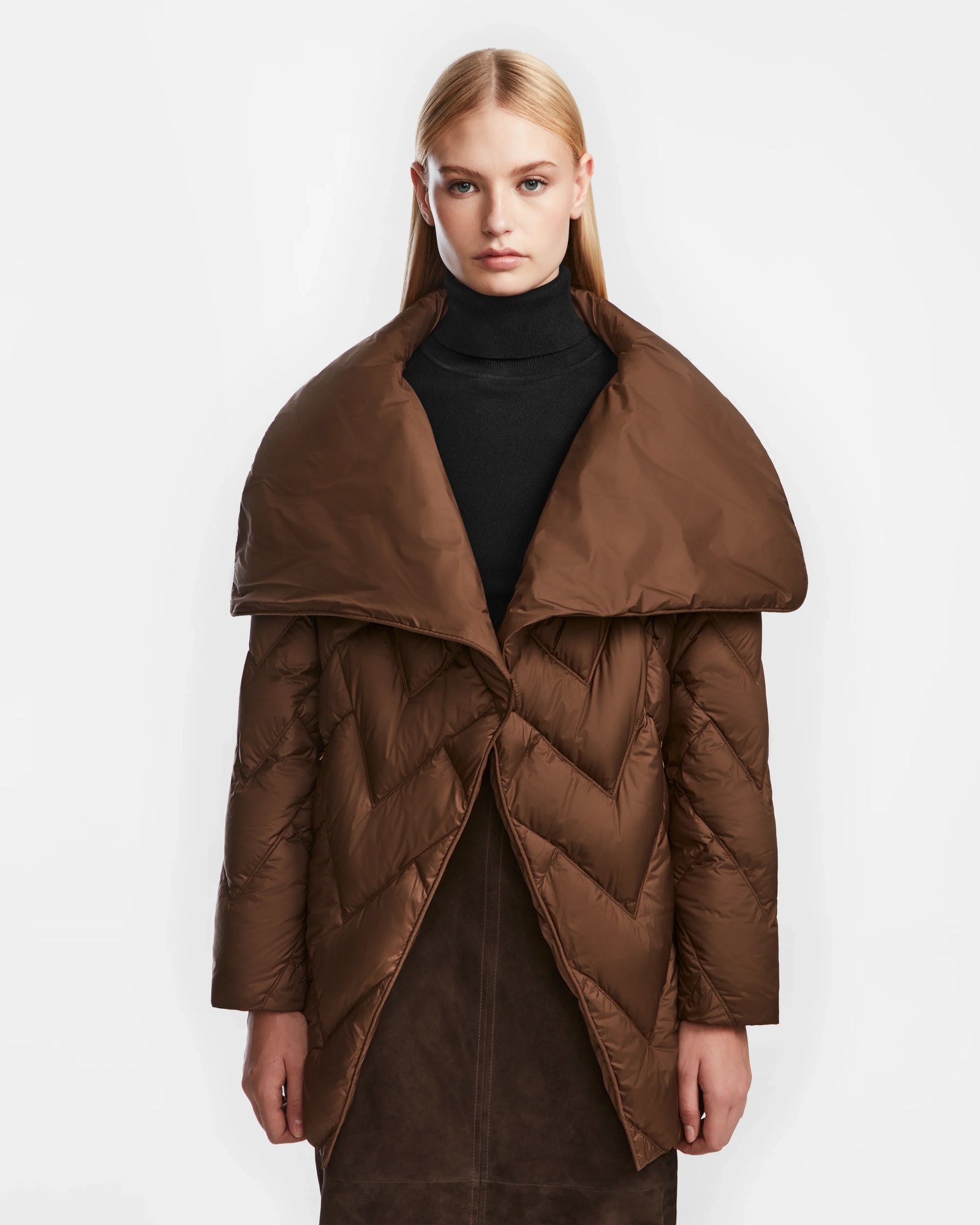 A woman with straight blonde hair wears the Rudsak ANNE MARIE - 8124713 Mocha, a womens relaxed fit brown puffer jacket with a wide collar, over a black turtleneck. She stands facing forward against a plain white background.