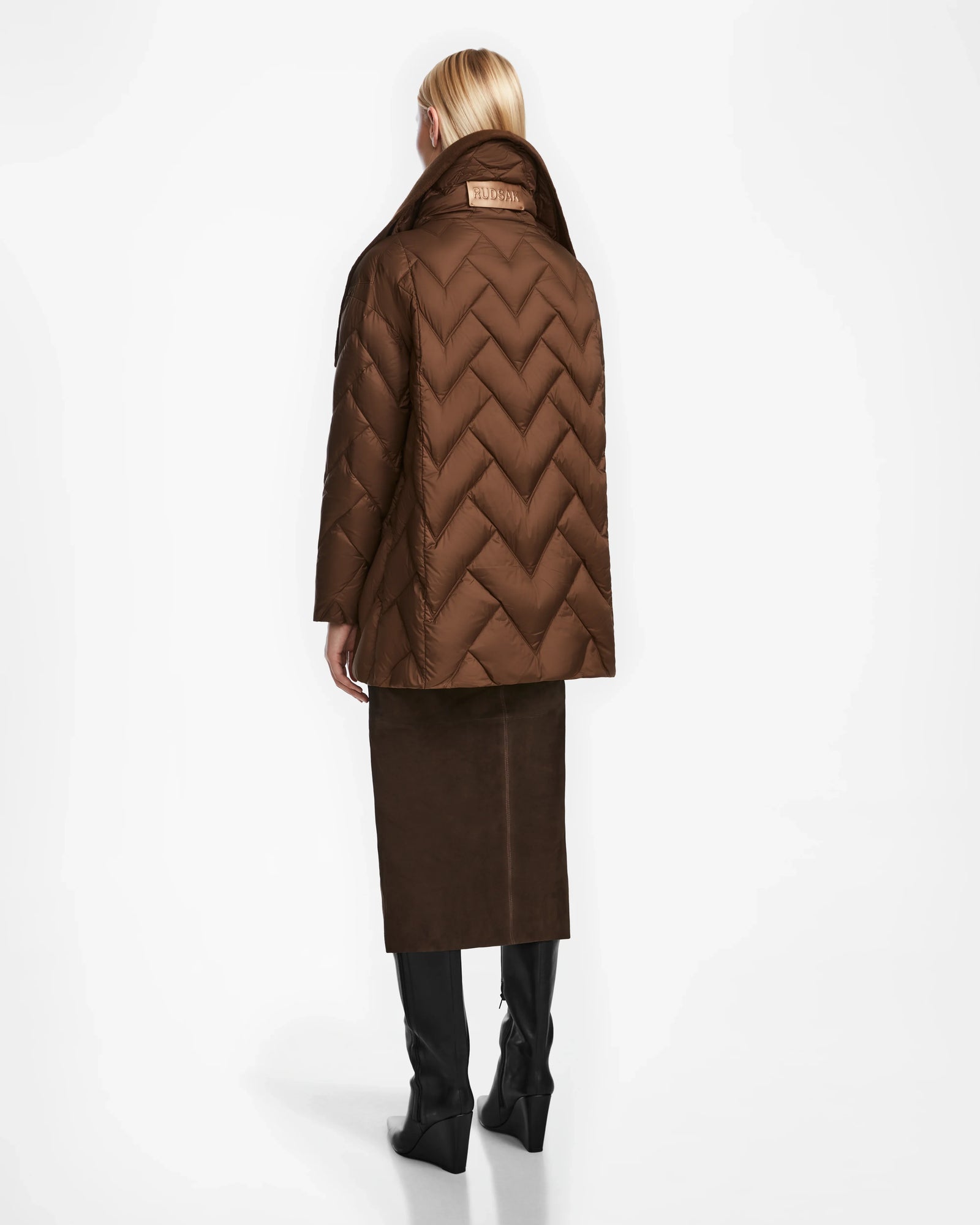 A woman with long blonde hair, seen from behind, wears the Rudsak ANNE MARIE - 8124713 Mocha brown quilted down wool jacket, paired with a brown knee-length skirt and black knee-high boots against a plain white background.