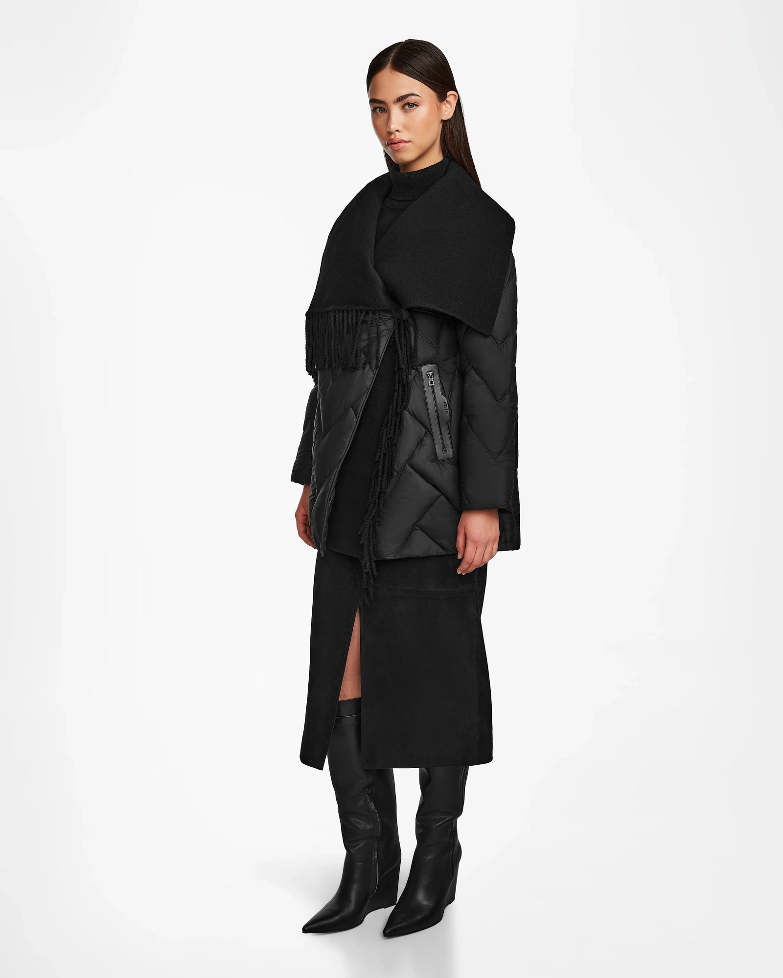 A woman stands against a white background wearing the Rudsak ANNE MARIE - 8124713 Black quilted down wool jacket with large lapels, paired with a black turtleneck, black front-slit midi skirt, and black knee-high boots.