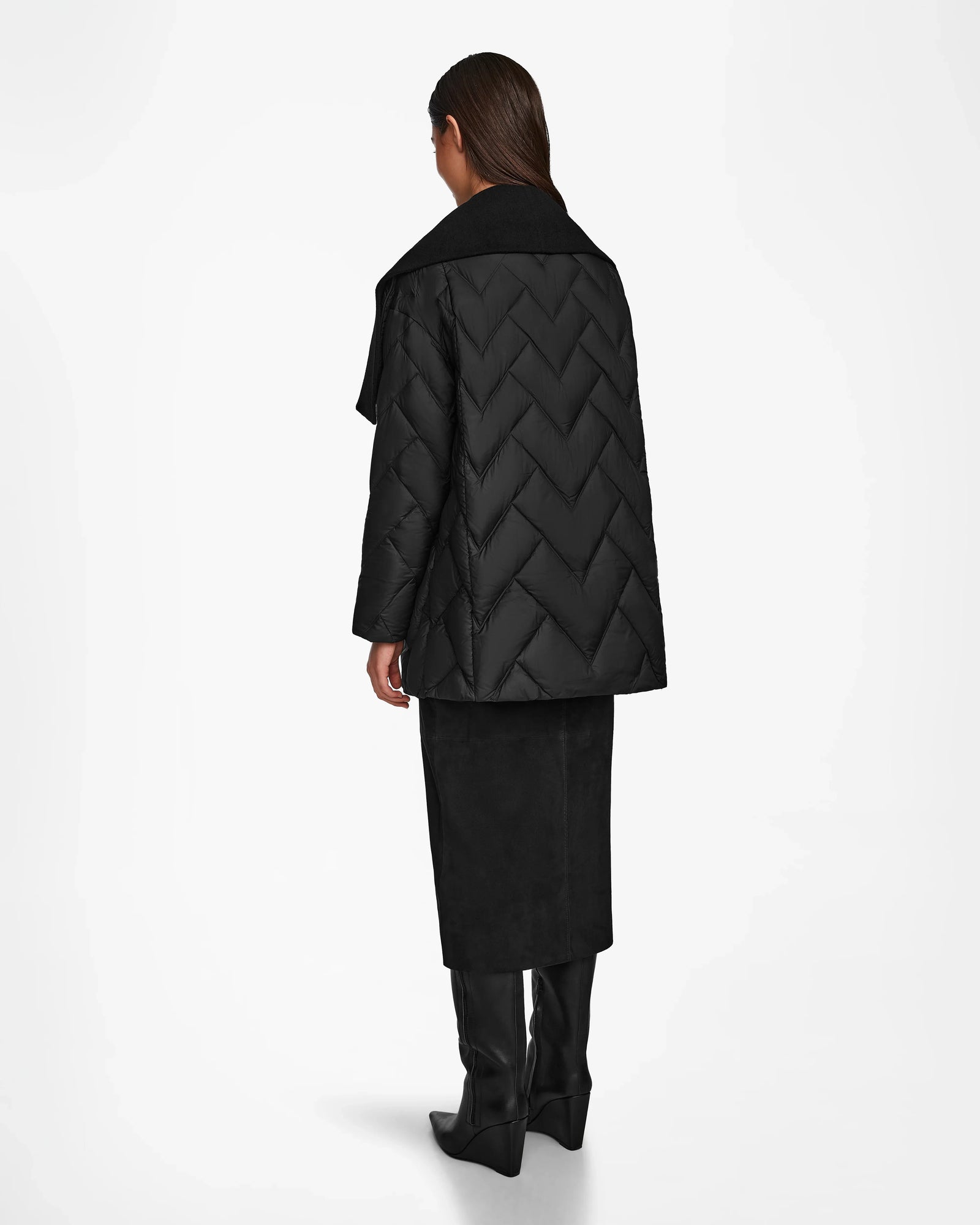 A woman with long dark hair faces away, wearing the Rudsak ANNE MARIE - 8124713 Black mid-length quilted down wool jacket, a black skirt, and black knee-high boots against a plain white background.