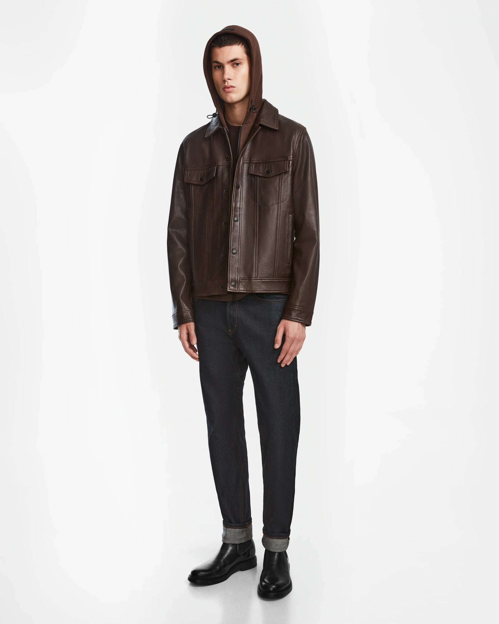 A person stands against a white background wearing the Rudsak AREN - 6124580 Espresso mens leather shirt jacket over a brown hoodie, paired with dark cuffed jeans and black boots.