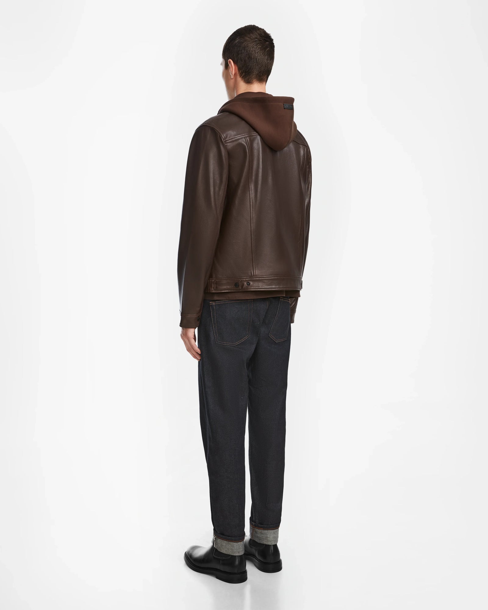 A man faces away, wearing the Rudsak AREN - 6124580 Espresso jacket over a brown hoodie, paired with dark cuffed jeans and black shoes against a plain white background.