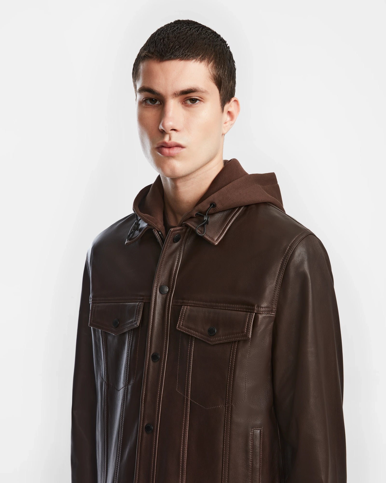 A young man with short dark hair wears a Rudsak AREN - 6124580 Espresso mens leather shirt jacket over a brown hoodie, standing against a plain white background.
