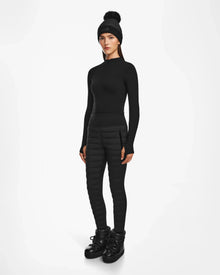 A woman faces forward in the Rudsak ARIL M - 8522514 BLACK fitted long-sleeve top, quilted pants, winter boots, and pompom hat—an effortlessly après-ski chic look against a white background.