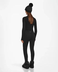 A woman faces away in apres-ski chic, wearing the Rudsak ARIL M - 8522514 BLACK long-sleeve top and sustainable insulated pants with boots and a pom-pom beanie. Her straight brown hair stands out against the white background.