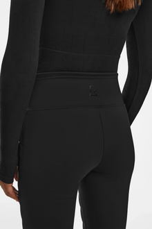 A person wears a fitted black long-sleeve top and high-waisted Rudsak ARIL M - 8522514 BLACK sustainable insulated pants, shown from the back. The pants have a small stitched logo on the waistband for an après-ski chic look.