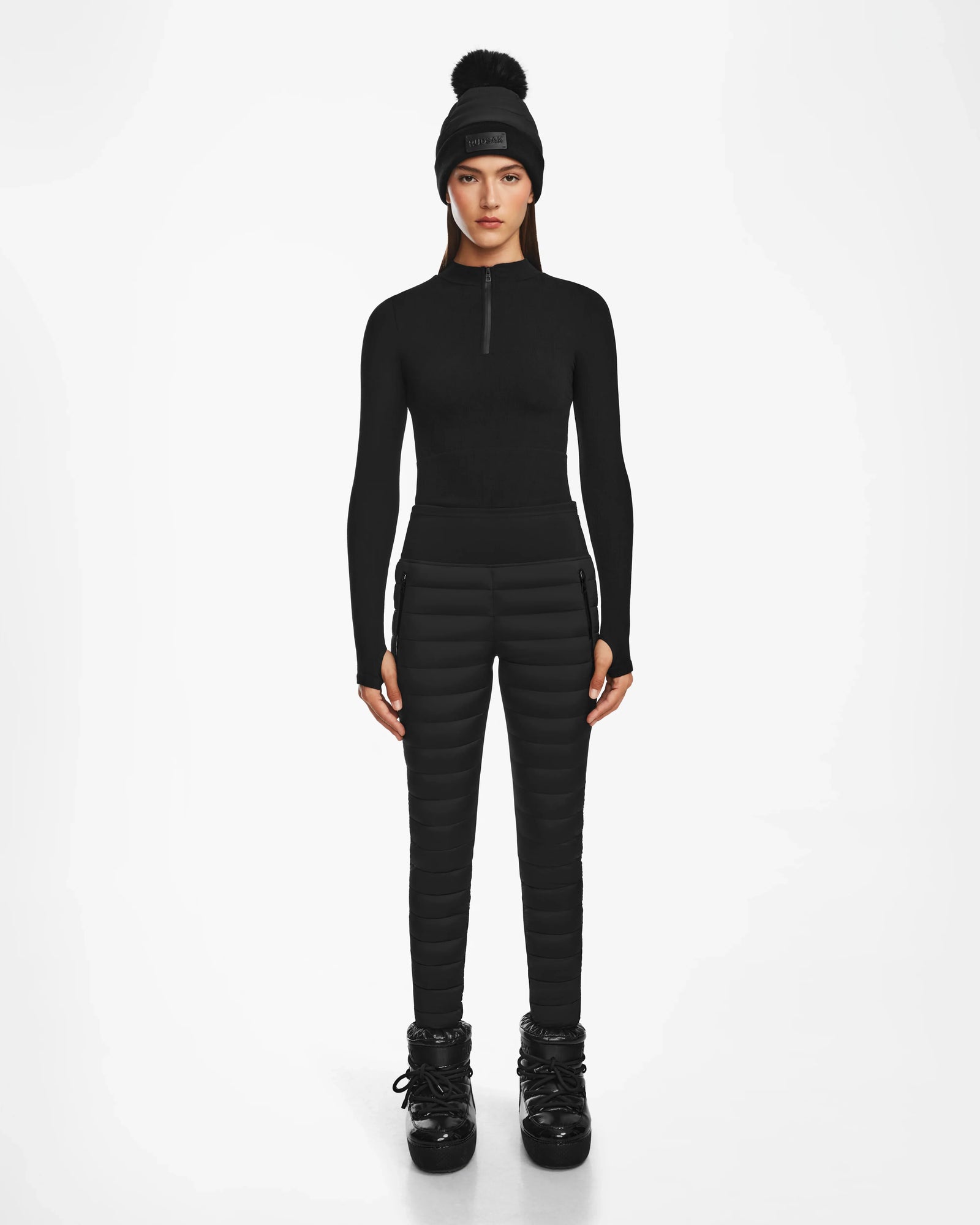 A woman faces forward against a white background, wearing the Rudsak ARIL M - 8522514 BLACK: a chic black fitted long-sleeve top paired with quilted pants, a pom-pom beanie, and winter boots for an après-ski look.