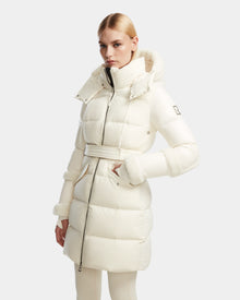 A woman wears the Rudsak AUDREY L - 8124585 Iceland knee-length down puffer, made from recycled Stormshell fabric with a high collar and fur-trimmed hood and cuffs, standing against a plain light background.