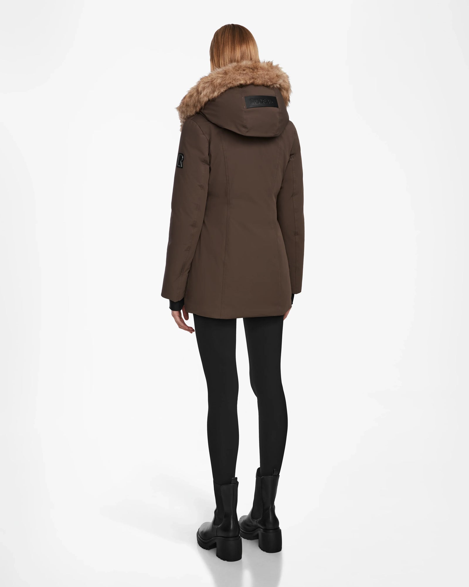 A person with straight, shoulder-length hair stands with their back to the camera wearing the Rudsak AURORE - L123561 Espresso women’s down parka with faux fur trim, black leggings, and black boots against a white background.