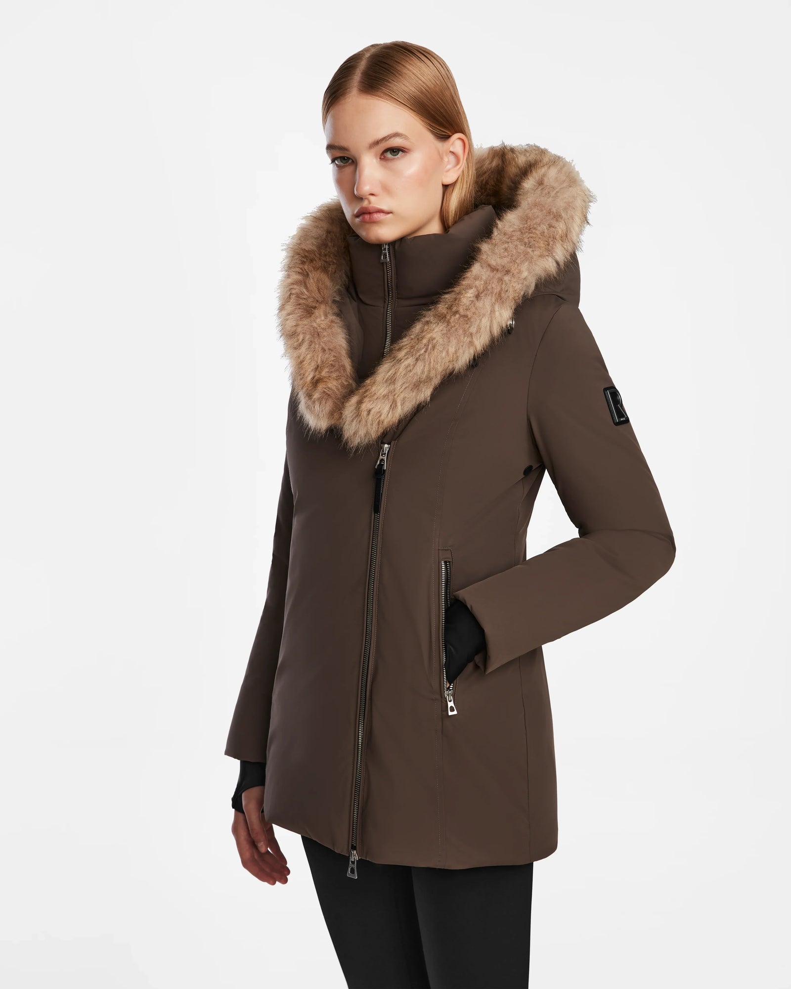A woman stands slightly turned against a white background wearing the Rudsak AURORE - L123561 Espresso, a fitted down parka with a large, fluffy fur-lined hood.