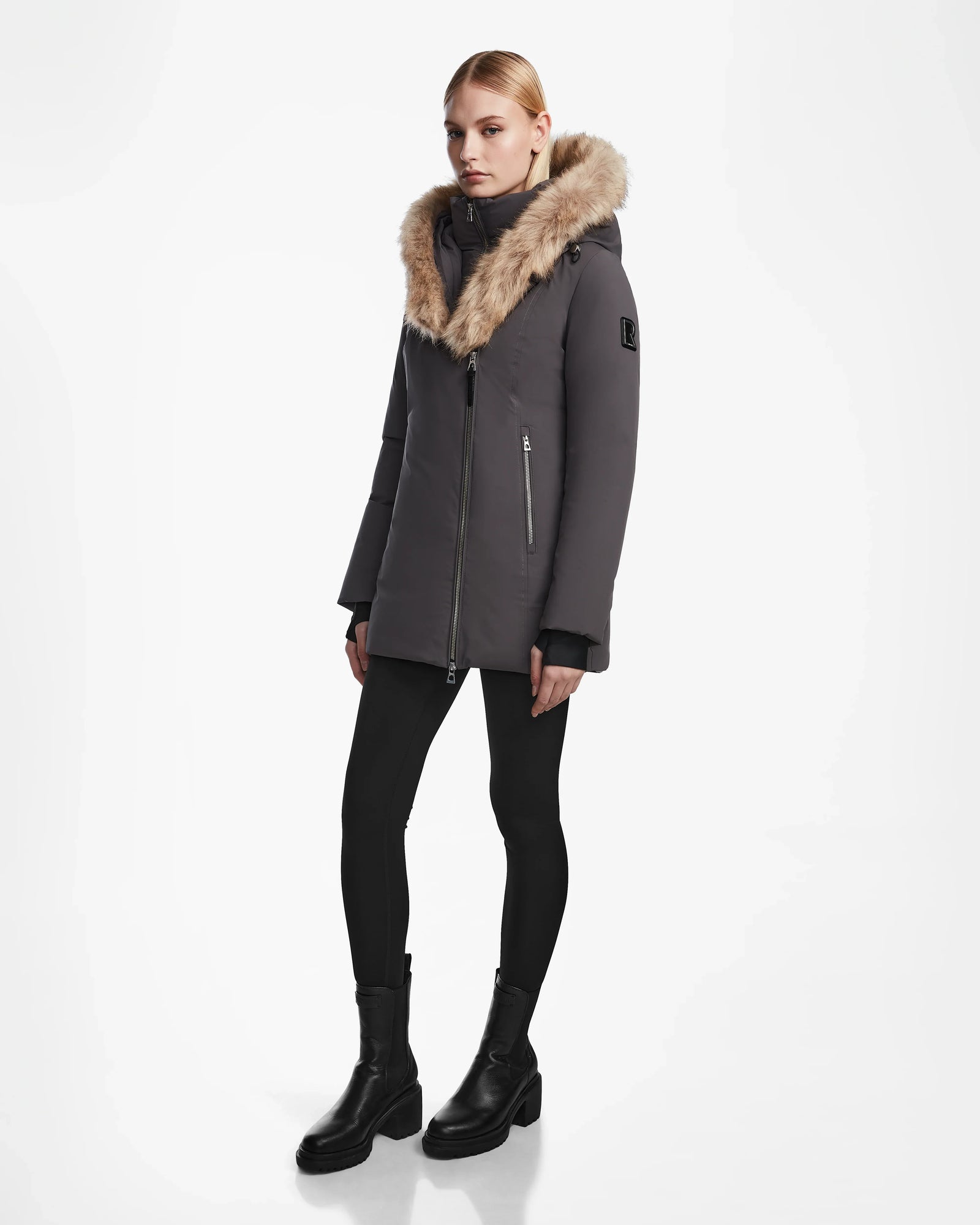 A woman wears the Rudsak AURORE - L123561 Grey fitted winter coat with a fur-lined hood, paired with black leggings and ankle boots, set against a plain white background.