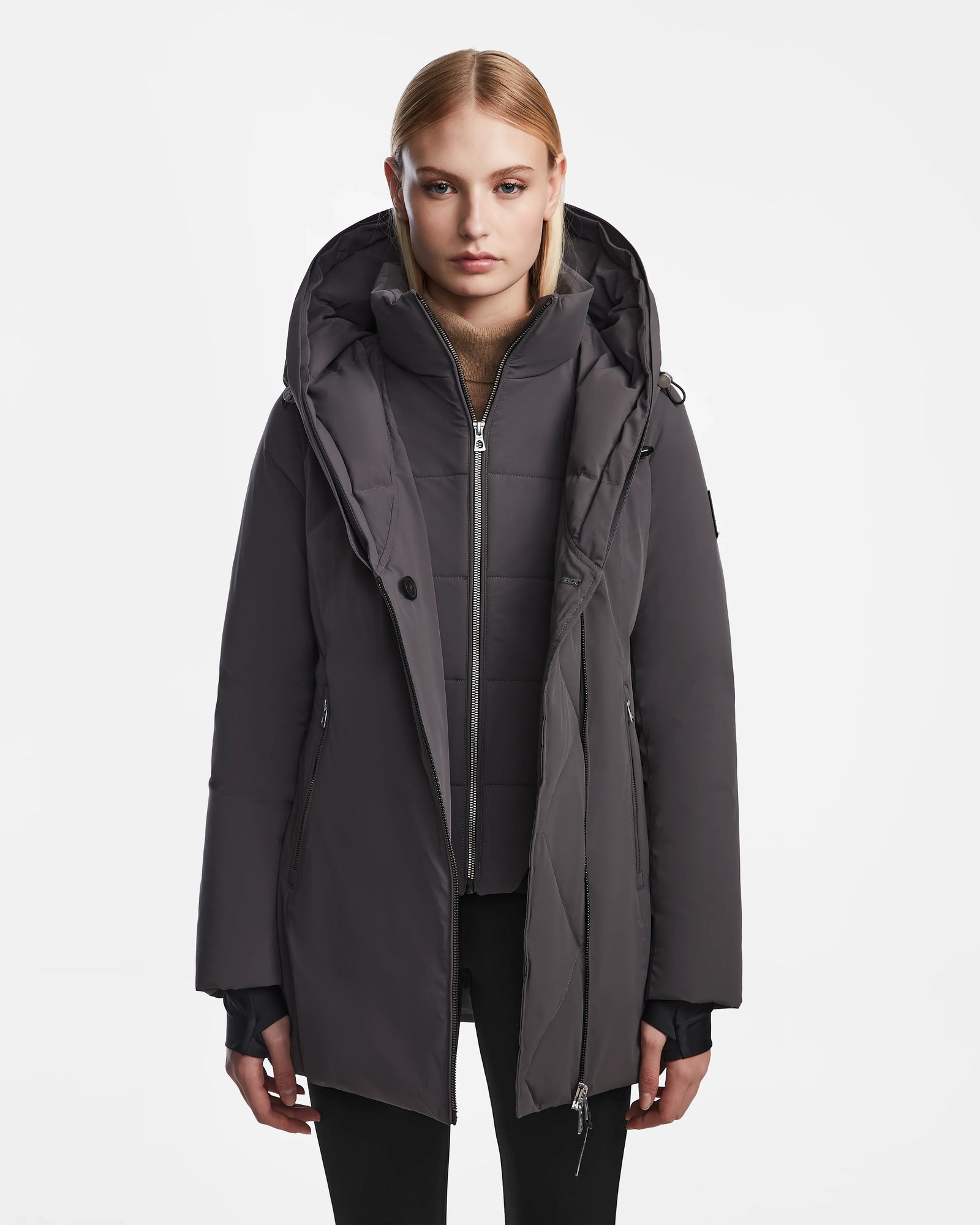 A woman with straight blonde hair wears the Rudsak AURORE - L123561 winter coat in grey, featuring a fitted silhouette and hood, paired with black pants. She stands against a white background, looking directly at the camera with a neutral expression.