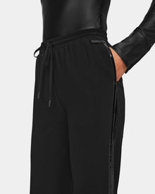 A person wearing a shiny black long-sleeve top and Rudsak CARDI - 8524561 Black womens pull-on trousers, featuring a relaxed fit and subtle side stripe detail, with hands in the pockets.